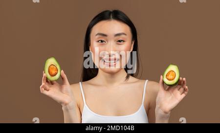 Organische Hautpflege. Eine glückliche asiatische Dame, die Avocado-Hälften in den Händen hält und ätherische Öle für Schönheitsbehandlungen genießt Stockfoto