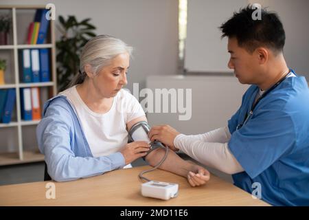 Ein seriöser, reifer koreanischer Arzt misst den Druck einer alten europäischen Patientin in der Klinik mit dem Tonometer Stockfoto