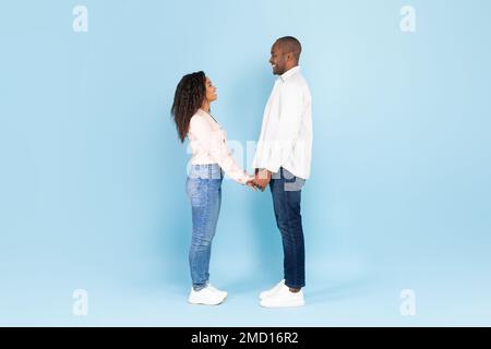 Die ganze Länge eines schwarzen Paares, das Hände hält und sich vor blauem Hintergrund anschaut, Seitenansicht, freier Raum Stockfoto