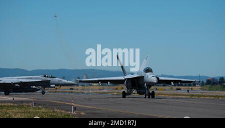 Eine F-18 Super Hornet aus dem Streikkämpfer-Geschwader der Navy (VFA) 192 „Golden Dragons“ aus dem Luftwaffenstützpunkt Lemoore, Kalifornien, fährt mit dem Taxi auf die Landebahn am Portland Air National Guard Base (PANGB), Oregon. Am 12. Juli 2022 startet ein F-15 Eagle mit 142. Flügeln im Hintergrund. Die F-15 Eagle-Piloten von PANGB nehmen am zweiwöchigen Dissimilar Air Combat Training (DACT) mit den Golden Dragons vom 11. Bis 22. Juli Teil. Stockfoto