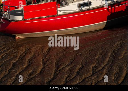 Am späten Abend spiegelt der dunkelbraune Schlamm, der das Bett des Flusses Tillingham auskleidet, ein rötliches Licht vom bemalten Rumpf einer Yacht wider, die bei Ebbe am Strand Quay, unterhalb der historischen Stadt und dem ehemaligen mittelalterlichen Cinque Port of Rye in East Sussex, England, festgemacht wurde. Das Tillingham fließt in den Fluss Rother, Ryes wichtigstes Outlet zum Ärmelkanal. Stockfoto