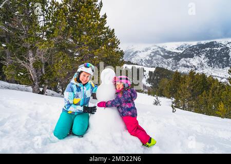 Familie, Mutter und Tochter, umarmter Schneemann mit Wald und Bergkette im Hintergrund. Skiurlaub im Winter in Andorra, Pyrenäen Stockfoto