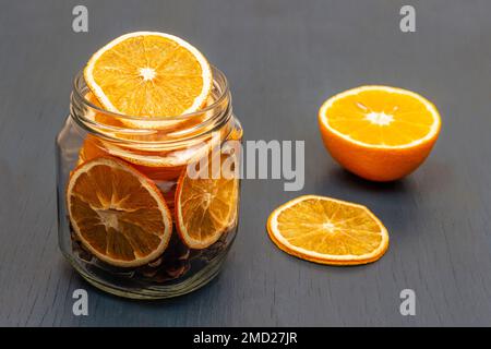 Eine halbe frische Orange auf dem Tisch. Trocknen Sie die orangefarbenen Scheiben und Tannenzapfen in einem Glas. Grauer Hintergrund. Stockfoto