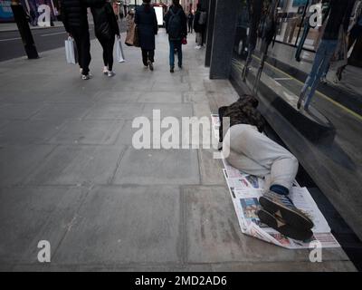 Ein Obdachloser, der in einem Zeitungsladen in der Oxford Street, London, schläft, während die Einkäufer mit Einkaufstaschen vorbeilaufen Stockfoto