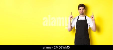 Unzufriedener und skeptischer Kellner mit schwarzer Schürze, der die Finger nach oben zeigt, drückt Abneigung und Gleichgültigkeit aus und steht auf gelbem Hintergrund Stockfoto