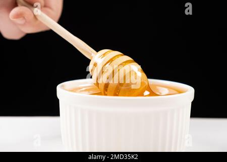 Honig in einer weißen Keramikschüssel mit einem Holzlöffel auf schwarzem Hintergrund. Stockfoto