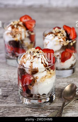 Kalter Sommerdessert - Plombir-Eiskrem mit Schokoladensoße, Kekskrümeln, reifen Erdbeeren und zerdrückten Erdnüssen in einem Glas auf einem Holztisch Stockfoto