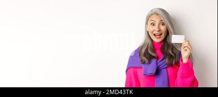 Einkaufskonzept. Glücklich asiatische alte aldy suchen beeindruckt und zeigt Kunststoff-Kreditkarte ihrer Bank, über weißem Hintergrund stehen Stockfoto