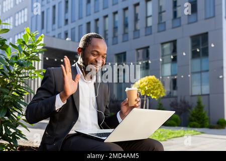 afroamerikanischer Geschäftsmann außerhalb des Bürogebäudes, der sich mit Kollegen aus der Ferne unterhält, Chef im Geschäftsanzug mit Laptop, der Videoanrufe führt, lächelt mit Kopfhörern, die auf der Bank sitzen. Stockfoto