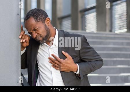 Herzinfarkt, afroamerikanischer Mann hat starke Brustschmerzen, Geschäftsmann außerhalb des Bürogebäudes, auf Treppen, die sich mit Brustschmerzen wickeln. Stockfoto