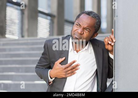 Herzinfarkt, afroamerikanischer Mann hat starke Brustschmerzen, Geschäftsmann außerhalb des Bürogebäudes, auf Treppen, die sich mit Brustschmerzen wickeln. Stockfoto