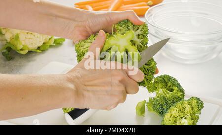 Die Frau schneidet Brokkoli auf ein weißes Schneidebrett. Frisches rohes Bio-Gemüse aus der Nähe auf dem Küchentisch. Gemüsesuppe oder Salatrezept Stockfoto