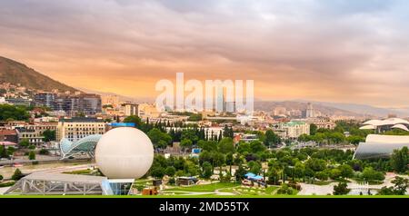 Tiflis, Georgia, mit Rike Park am Ufer des Mtkvari oder der Kura, Friedensbrücke, Heißluftballon und Konzertsaal bei Sonnenuntergang mit dramatischem Himmel Stockfoto