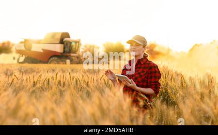 Frau Farmer mit digitalem Tablet auf einem Hintergrund von Harvester. Smart Farming-Konzept. Stockfoto