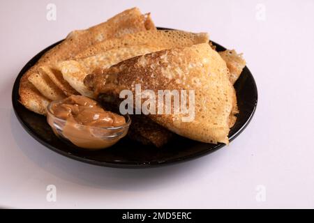 Frittierte, leckere Pfannkuchen und abgekochte Kondensmilch in einer kleinen Schüssel, die auf einem Teller auf weißem Hintergrund liegt Stockfoto