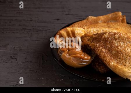 Dünne Pfannkuchen mit abgekochter Kondensmilch liegen wunderbar auf einem Teller auf der Seite vor dunklem Hintergrund Stockfoto