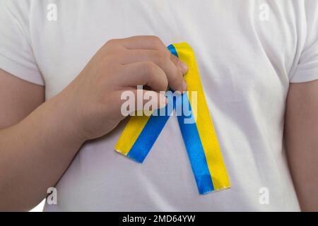 Mädchen in einem weißen T-Shirt mit einer Schlaufe aus gelb-blauem Textilband (Nahaufnahme). Staatsflagge der Ukraine. Nationales Symbol der Ukraine. Patriot Stockfoto