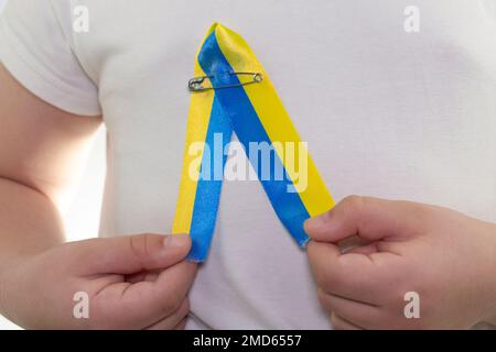 Mädchen in einem weißen T-Shirt mit einer Schlaufe aus gelb-blauem Textilband (Nahaufnahme). Staatsflagge der Ukraine. Nationales Symbol der Ukraine. Patriot Stockfoto