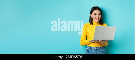 Glückliche asiatische Frau in gelben Pullover mit Laptop, online einkaufen oder arbeiten, über blauen Hintergrund stehen Stockfoto