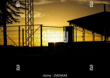 Kontrastierende Silhouette eines Industriegebäudes hinter Gitterzaun vor dem gelben Abendhimmel bei Sonnenuntergang. Lagerhaus, Dach des Fabrikgebäudes. Kommerziell Stockfoto