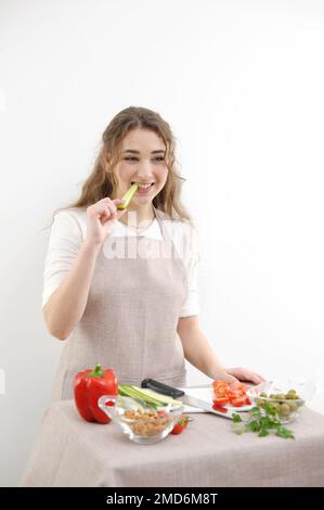 Junge Frau Koch beißt Gurke bereitet geschnittenes Gemüse oder Salat sie ist in beige Küchenschürze auf weißem Hintergrund gesundes Essen Konzept vegetarisches Essen Ja Schönheit und Jugend gibt Platz für Text Stockfoto