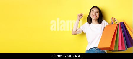 Glückliche asiatische Frau Gefühl zufrieden nach dem Einkaufen im Verkauf, halten Papiertüten und Stretching mit glücklichen Lächeln, stehend über gelbem Hintergrund Stockfoto