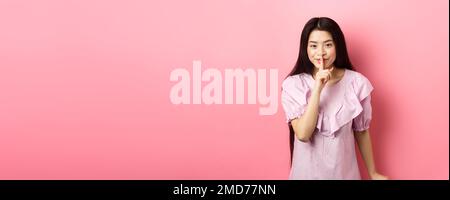 Nettes asiatisches Mädchen versteckt Geheimnis, mit Finger auf die Lippen gedrückt und lächelnd, bitten, ruhig zu bleiben, im Kleid auf rosa Hintergrund stehen Stockfoto