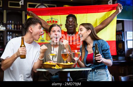 Begeisterte, vielfältige Sportfans mit spanischer Flagge, die ein Gewinnerspiel mit Gläsern Bier und Chips in der Kneipe feiern Stockfoto