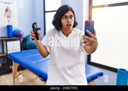 Junge hispanische Physiotherapeutin Frau, die Handgriff hält, um Muskeln zu trainieren, Videoanrufe im Schockgesicht macht, skeptisch und sarkastisch wirkt, überrascht Stockfoto