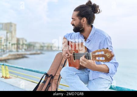 Junger hispanischer Musiker, der klassische Gitarre spielt und auf der Balustrade am Meer sitzt Stockfoto