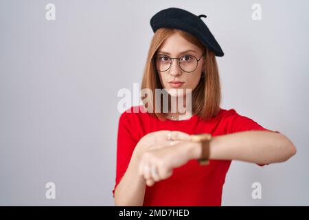 Junge rothaarige Frau mit Brille und Baskenmütze in Eile, zeigt auf die Uhrzeit, Ungeduld, aufgebracht und wütend wegen Deadline-Verzögerung Stockfoto