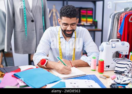 Ein junger arabischer Schneider, der selbstbewusst in einem Schneiderladen auf ein Notebook lächelt Stockfoto