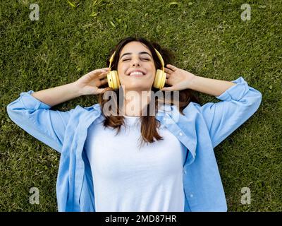 Junge, fröhliche Millennials, die auf dem Rasen liegt und Musik mit Kopfhörern hört Stockfoto