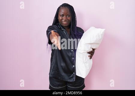 Junge afrikanische Frau mit Pijama-Kissen, lächelnd und fröhlich, mit Handfläche, die Hilfe und Akzeptanz bietet. Stockfoto