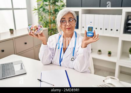 Eine Frau mittleren Alters mit grauen Haaren, die eine Arztuniform trägt und das Glukoseüberwachungsgerät hält, die zur Seite lächelt und wegstarrt und denkt. Stockfoto