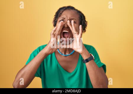 Afrikanische Frau mit Dreadlocks, die auf gelbem Hintergrund steht und laut schreit, mit den Händen über dem Mund Stockfoto