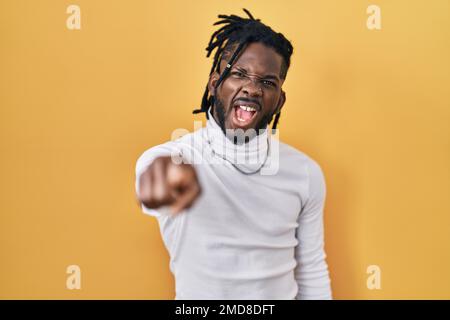 Afrikanischer Mann mit Dreadlocks, der einen Rollkragenpullover auf gelbem Hintergrund trägt und unzufrieden und frustriert auf die Kamera zeigt, wütend und wütend auf die Kamera Stockfoto