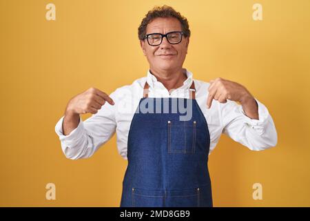 ein hispanischer Mann im mittleren Alter, der eine professionelle Kochschürze trägt, selbstbewusst aussieht und mit einem Lächeln im Gesicht auf sich zeigt, stolz und glücklich mit den Fingern. Stockfoto