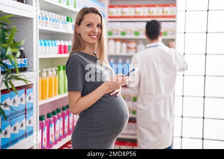 Mann und Frau, Apotheker und schwangere Klientin, die in der Apotheke ein Smartphone benutzen Stockfoto