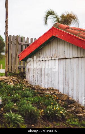 Vertikaler Blick auf ein bescheidenes Holzhaus, weiß gestrichen mit orange-roten Fliesen in A-Form, gelegen an einem kleinen Steinhügel mit Unterholz, eingezäunt Stockfoto