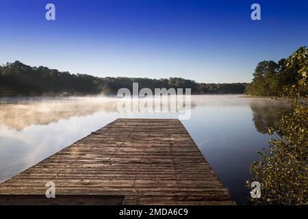 Ein hölzerner Dock erstreckt sich über einen ruhigen See an einem nebeligen Tag. Stockfoto