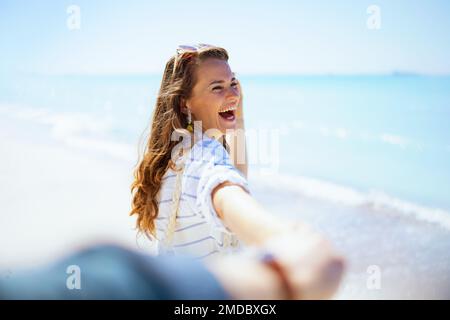 Eine lächelnde elegante 40-jährige Frau am Strand in einem weißen gestreiften Hemd und Shorts, die Spaß haben. Stockfoto