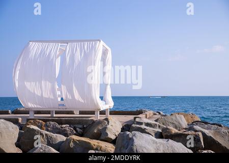 Malerischer Blick leere weiße Strand-Hütten aus Holz mit weißen Vorhängen, die vom Wind gegen das Meer oder das Meer an der san-Küste wehen. Luxuriöse Sonnenliege für Spa und Stockfoto