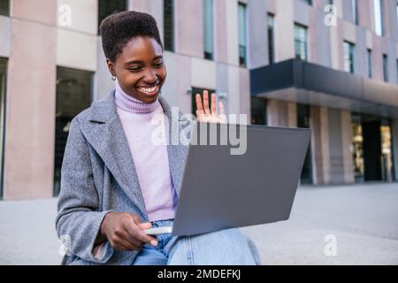 Fröhliche afroamerikanische Freiberuflerin in stilvoller Oberbekleidung, die lächelt und winkt. Sie sitzt auf einer Bank und führt Videoanrufe über einen Laptop auf der Straße der Stadt Stockfoto