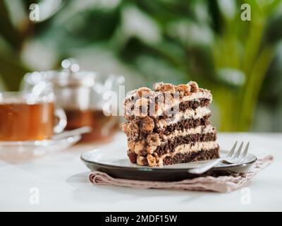 Perfekter ästhetischer Schichtenkuchen mit Karamell und Nüssen auf dem Teller über dem grauen Tisch. Stück Schokoladenmandelkeks mit Karamell-Zuckerguss in der Nähe von Teetasse und Teekannen. Stück Biskuitkuchen im Café Stockfoto