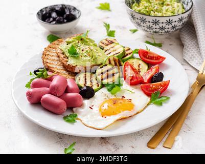 Spiegelei mit Mini-Würstchen, Tomaten, gegrillte Zucchini, schwarze Oliven und Toast mit Guacamole. Englisches Frühstück. Brunch. Stockfoto