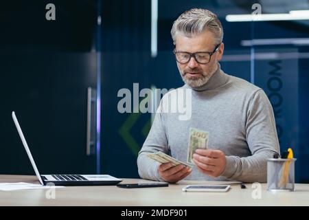 Ein seriöser, reifer Investor, der im Büro arbeitet, ein grauhaariger Geschäftsmann, der Bargeld zählt, ein Geschäftsmann, der in einem modernen Büro mit einem Laptop arbeitet. Stockfoto