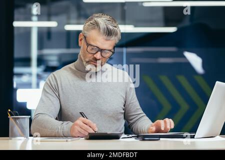 Ein seriöser Geschäftsmann im Büro, der mit einem Laptop arbeitet, ein grauhaariger Senior-Mann, der am Schreibtisch sitzt. Stockfoto