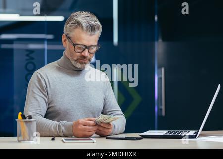 Ein seriöser, reifer Investor, der im Büro arbeitet, ein grauhaariger Geschäftsmann, der Bargeld zählt, ein Geschäftsmann, der in einem modernen Büro mit einem Laptop arbeitet. Stockfoto