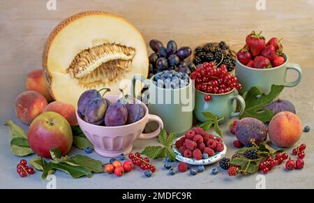 Verschiedene Sommerbeeren - Himbeeren, Erdbeeren, Heidelbeeren, Kirschen, Johannisbeeren, Pflaumen - in Bechern auf einem Holztisch Stockfoto